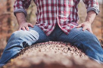 Strong man in plaid shirt and blue jeans sitting on a cut tree trunk, legs spread