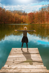 Fototapeta na wymiar Woman standing on the edge of wooden dock, with her arms raised up, looking at water and colorful autumnal forest
