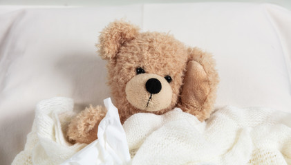 Cold, flu or allergy. Cute teddy in bed, covered with a warm blanket, holding a tissue