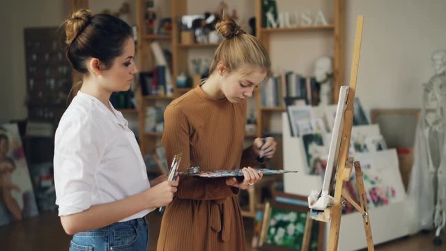 Young lady student is painting on canvas using oil paints while her teacher experienced artist is standing near her and looking at picture. Pictorial art and people concept.