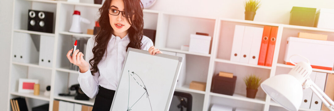 A beautiful young girl stands near an office desk and draws a magnetic marker on the magnetic board.