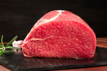 A photo of eye round beef, a raw cut, with rosemary, garlic, and pepper, on a black background with...