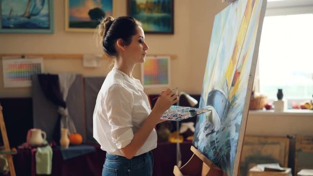 Pretty woman painter is depicting seascape then looking at her artwork and smiling enjoying beauty of picture. Satisfaction, creativity and people concept.