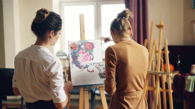 Back view of art student painting flowers on canvas and her helpful teacher standing nearby and checking her work. Visual arts and education concept.