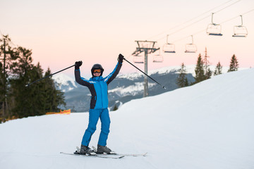 Girl enjoying ski holiday standing on the snowy mountain and raised her hands up. Woman at ski resort wearing helmet, blue ski suit and goggles.