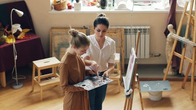 Good-looking young woman is holding palette and painting flowers under guidance of helpful teacher standing together in workroom of art school and talking.