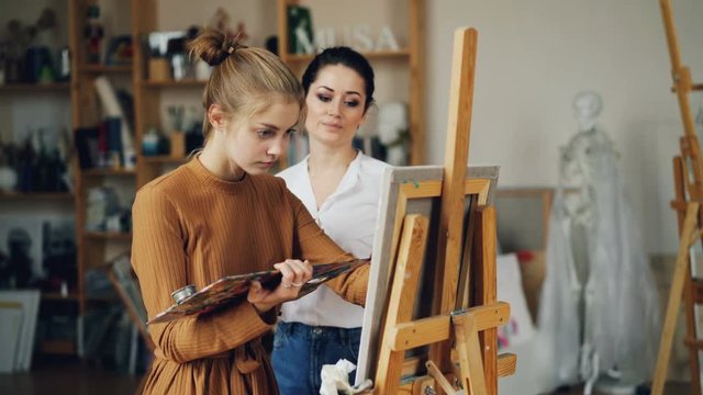 Experienced art teacher is working with pretty girl talanted student painting picture and talking sharing experience indoors in workroom full of artworks.