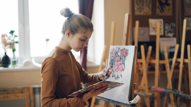 Diligent art school student is mixing bright colors on palette then painting flowers on canvas working indoors in cozy workroom with beautiful artworks.
