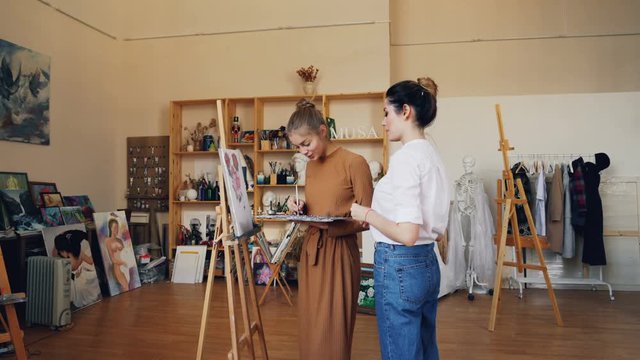 Cheerful lady is painting picture together with her female teacher professional artist using brush and oil paints. Modern workshop with easels and artworks is visible.