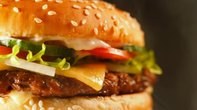 Juicy beef burger with cutlet, onion, vegetables, melted cheese, lettuce, sauce and topped sesame seeds. Isolated hamburger rotates on dark smoke background, close-up view.