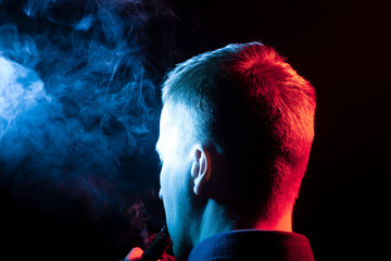 A view from the back of a man’s head around in a shirt smoking a vape and exhaling multi-colored smoke of blue to different sides from himself on a black isolated background.