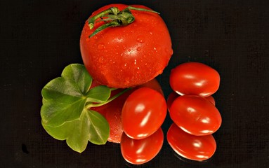 close up of a red tomato with three cherry tomatoes and a green leaf on a black background
