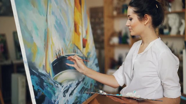 Smiling young woman is painting picture depicting boat and blue sea waves on canvas in oil paints, she is holding brush and palette standing near easel.