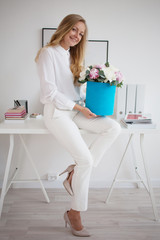 Stylish young blonde in office, white shirt and pants. Holds a luxurious bouquet of peonies in a blue hat box.