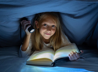 Pretty happy girl reading a book in darkness hiding under the duvet in bed with flash light