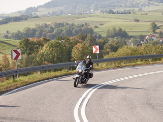 Obraz premium Bearded motorcyclist in helmet, sunglasses and black leather clothing driving bike along sharp turn of empty road on bright summer day on misty background of rural landscape and distant green hills.