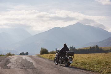 Back view of motorcycler in black leather jacket riding cruiser bike along road on blurred copy space background of beautiful foggy mountain range and blue cloudy sky on bright sunny summer day.