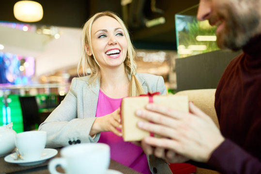 Portrait of adult couple exchanging presents at table in cafe, focus on happy woman holding gift box