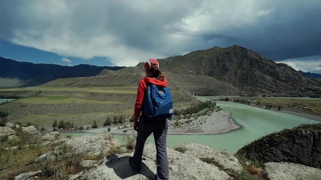 Young happy torist woman with backpack and cap reaches to the edge of the mountain and puts his hands in the air. Back view, slider