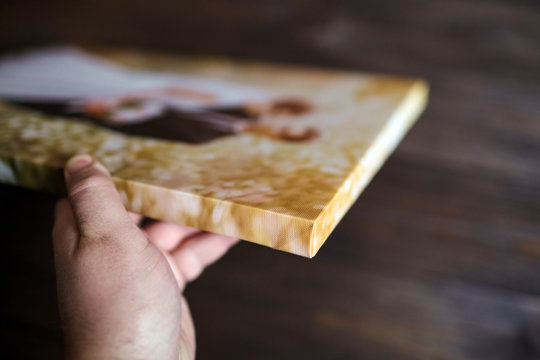 Male hand holding a photography with gallery stretch on a wooden frame. Printed wedding photo on canvas. Lateral side closeup. Selective focus