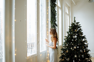 Christmas concert. Cute blonde woman in evening dress holding a glass of sparkling champagne on the background of a large beautifully decorated Christmas tree in the living room.