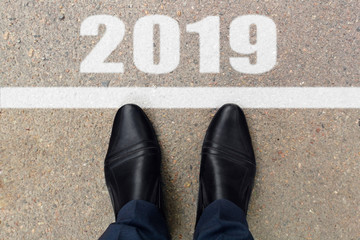 Man feet in black leather shoes standing before text 2019 painted on asphalt, top view