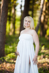 Portrait of beautiful pregnant woman in sheer long white maternity dress looking dreamy in the middle of forest