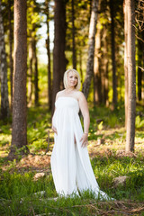 Obraz na płótnie Canvas Portrait of beautiful pregnant woman in sheer long white maternity dress looking dreamy in the middle of forest