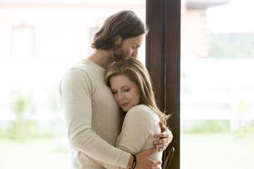 Sad young married couple embracing standing in living room opposite window at home. Sorrowful wife...