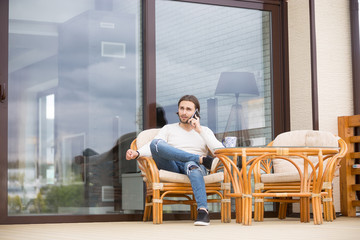 Young confident and serious millennial man sitting on outdoors armchair in terrace of his new...