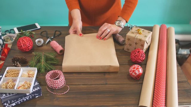 Female hands wrapping Christmas gift in craft ecological paper on dark wooden backround with decorating elements, colourfil twine stripes and pack paper rolls, Christmas or New year DIY packing.
