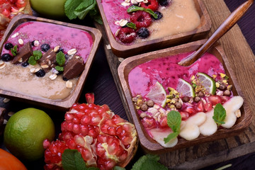 Different smoothie bowls topped with berries, fruits, nuts, oatmeal flakes, bran, chocolate and mint in wooden dishes surrounded by the ingredients