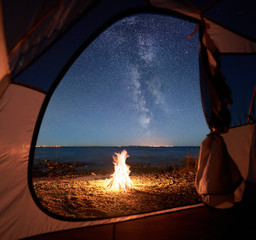 View from inside tourist tent. Night camping near seacoast. Burning campfire under beautiful night sky full of stars and Milky way