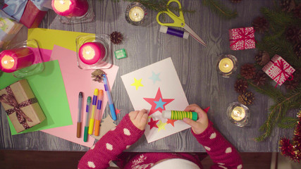 Top view. Little girl glues New Year card. Table with Christmas decorations.