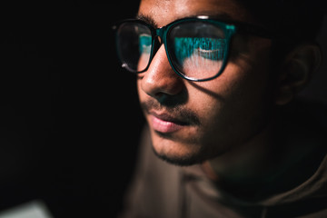Hacker in glasses and a hood works at a computer in the dark, a reflection in glasses close-up.