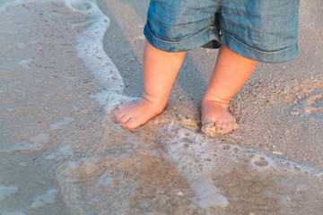 Fototapeta na wymiar Bare feet walking at sandy beach near the sea. Little baby in blue jeans shorts going to touch the sea at sunset. Wave washes baby's feet. Toned. Soft focus.