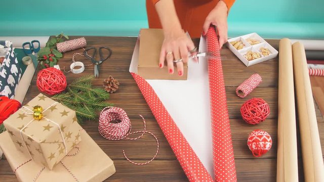 Woman in a red sweater wrapping gift box with equipment and decorating items on wood table, preparing for celebrating Christmas holidays - top view.