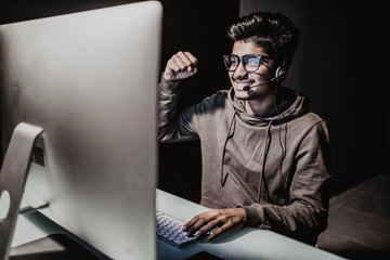 Ecstatic asian gamer boy rejoicing victory while playing video games on computer in dark room wearing headphones and using backlit keyboard © dianagrytsku