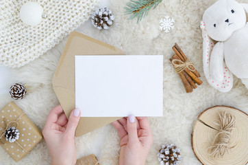 girl hands holds blank paper mock up with space for text, art work,lettering, pine cone fir branch,rope,rabbit toy on fur background. Flat lay, top view. Cosy, soft ,minimal. Christmas,new year