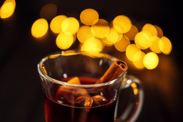 closeup piece of glass of mulled wine with orange and cinnamon on dark black background, on white plate, Christmas lights, large yellow bokeh