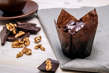 Chocolate muffin, standing on a white wooden surface and a napkin next to the pieces of chocolate and nuts.
