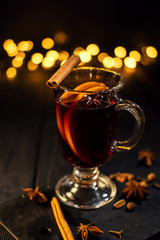 cinnamon lies in a glass, closeup glass of mulled wine with orange and cinnamon on dark black background,  Christmas tree and lights, large yellow bokeh, mulled wine set