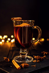 cinnamon lies in a glass, closeup glass of mulled wine with orange and cinnamon on dark black background,  Christmas tree and lights, large yellow bokeh, mulled wine set
