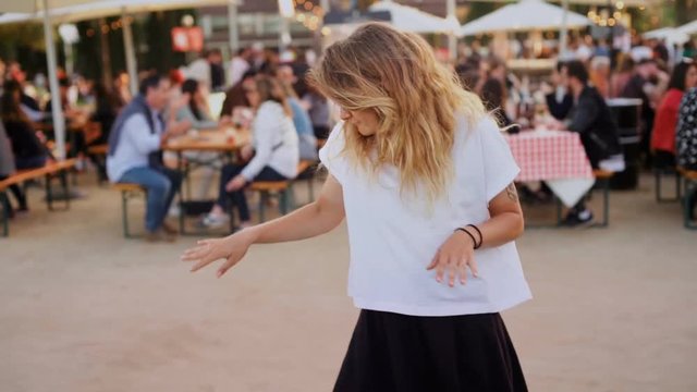 Happy and excited young female with blonde hair or hipster millennial dances funnily with humor, makes silly dance moves at party or festival in summer. Looks at camera and laughs, has good time