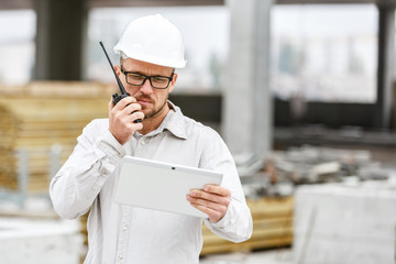 Male head engineer wearing white safety hardhat with walkie talkie and tablet inspecting...