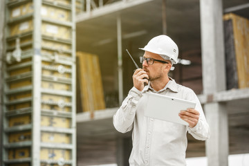 Male head engineer wearing white safety hardhat with walkie talkie and tablet inspecting construction site.