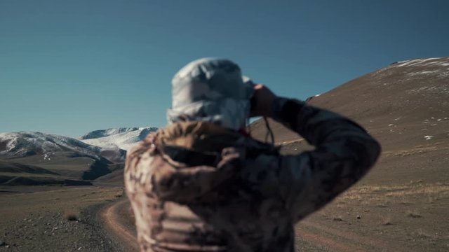 Man is watching mountain sheep through his binoculars. Study of argali, population. Snow peak and mountains in the background. Refocus. Back view