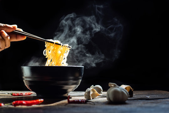 Hand uses chopsticks to pickup tasty noodles with steam and smoke in bowl on wooden background, selective focus. Asian meal on a table, junk food concept