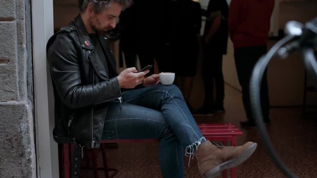 Casual middle aged adult man in black leather jacket, blue denim jeans and brown boots enjoys and drinks cup of coffee on open terrace of specialty cafe, scrolls through smartphone