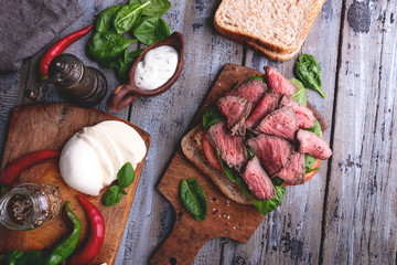 Steak sandwich, sliced roast beef, cheese,spinach leaves,tomato. Rustic,on a wooden surface
